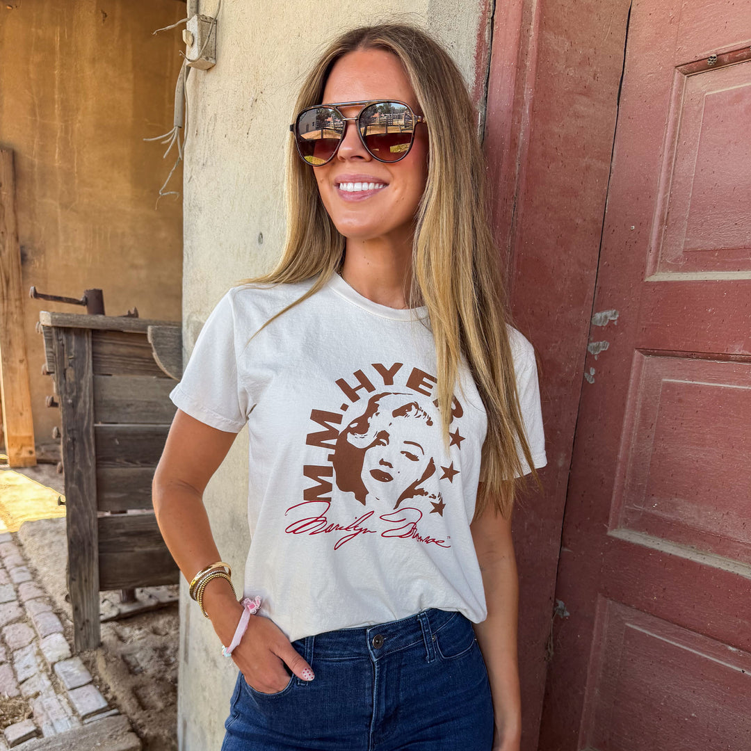 Woman wearing a white t-shirt with a graphic design, standing in front of a rustic wooden door.