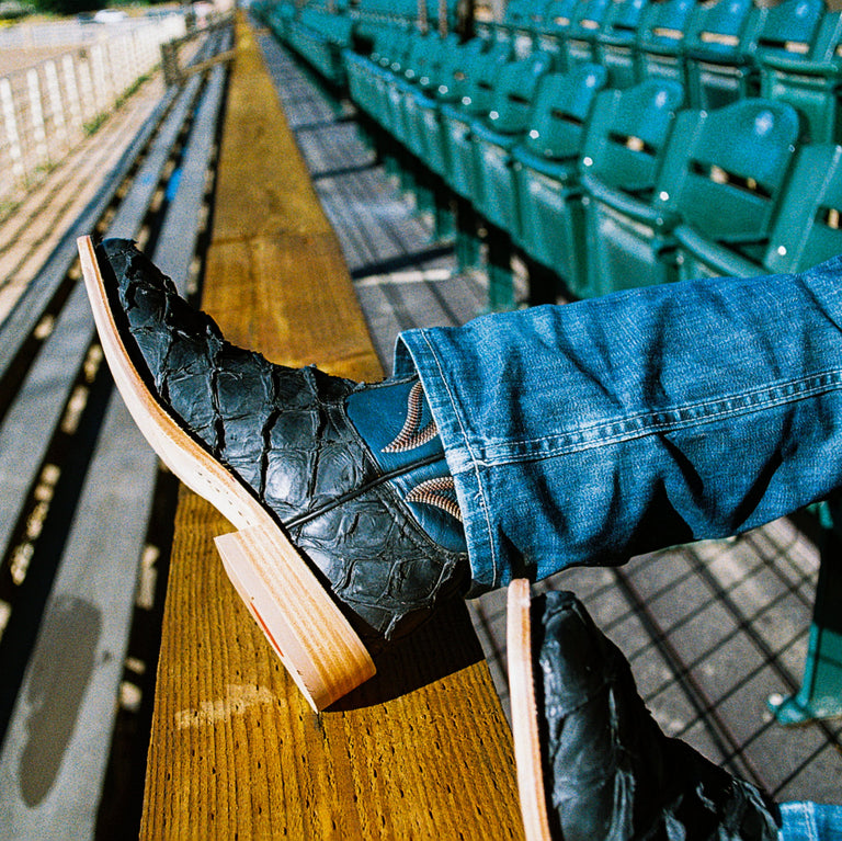 A leg resting on a wooden slat wearing a cowboy boot with black big bass scaled vamp and a blue vamp peeking out from under a pair of jeans