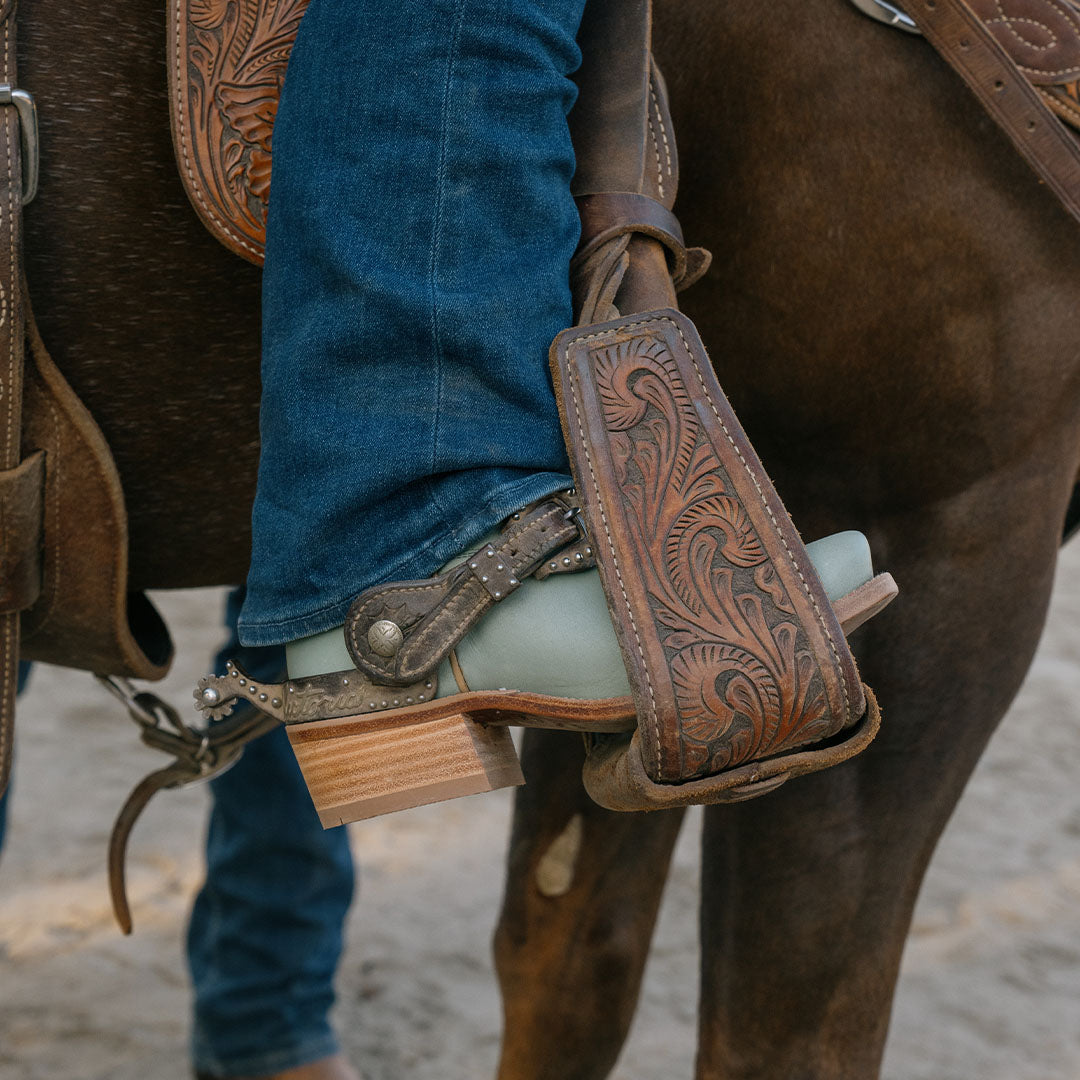 Lifestyle shot of a person wearing blue jeans and light blue cowboy boots with spurs in a stirrup on a horse