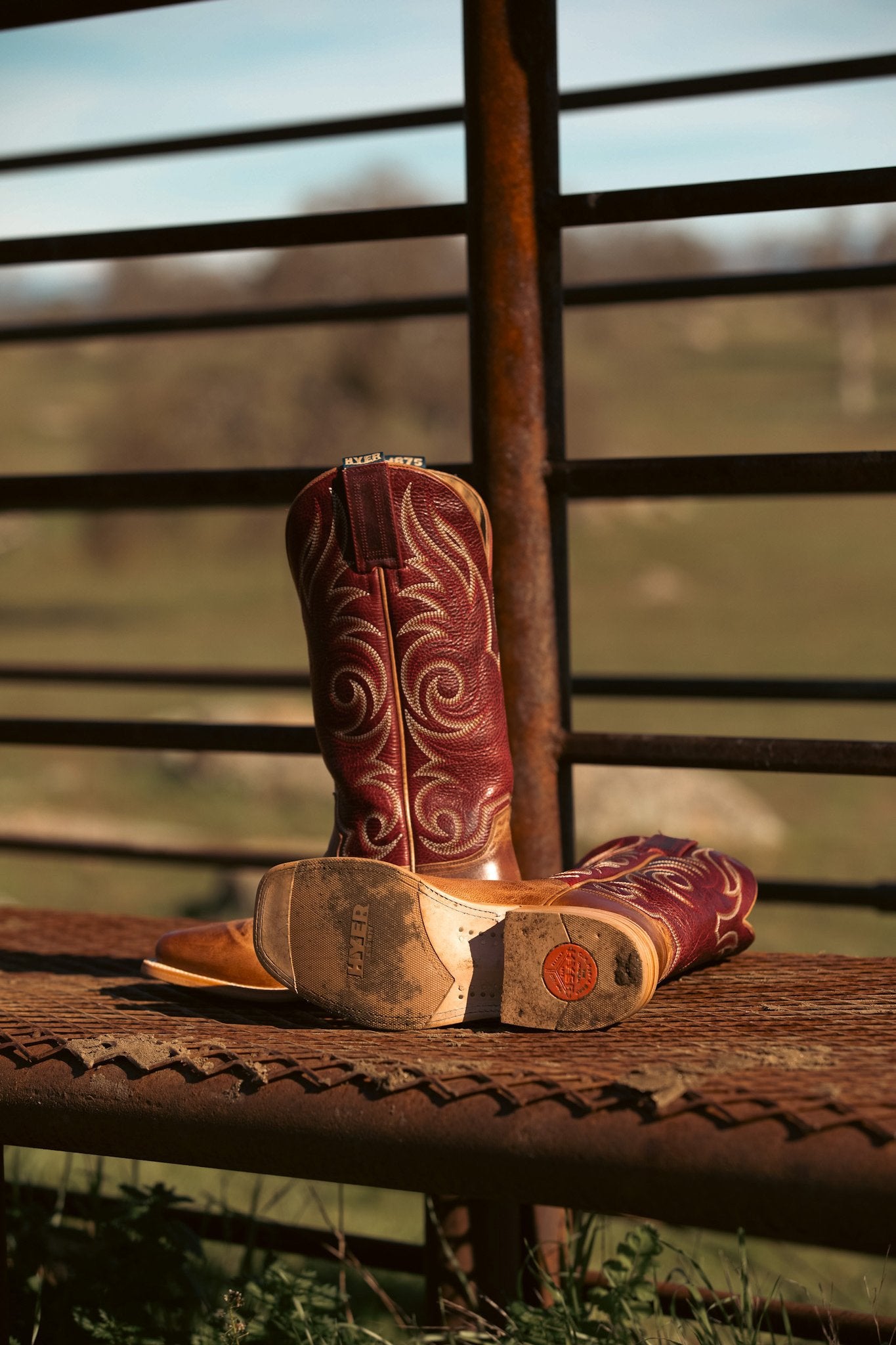 A pair of western cowboy boots with reddish-brown shafts and tan toes, featuring white embroidered swirl patterns, resting on a rustic metal bench in front of a fence with a blurred outdoor background.