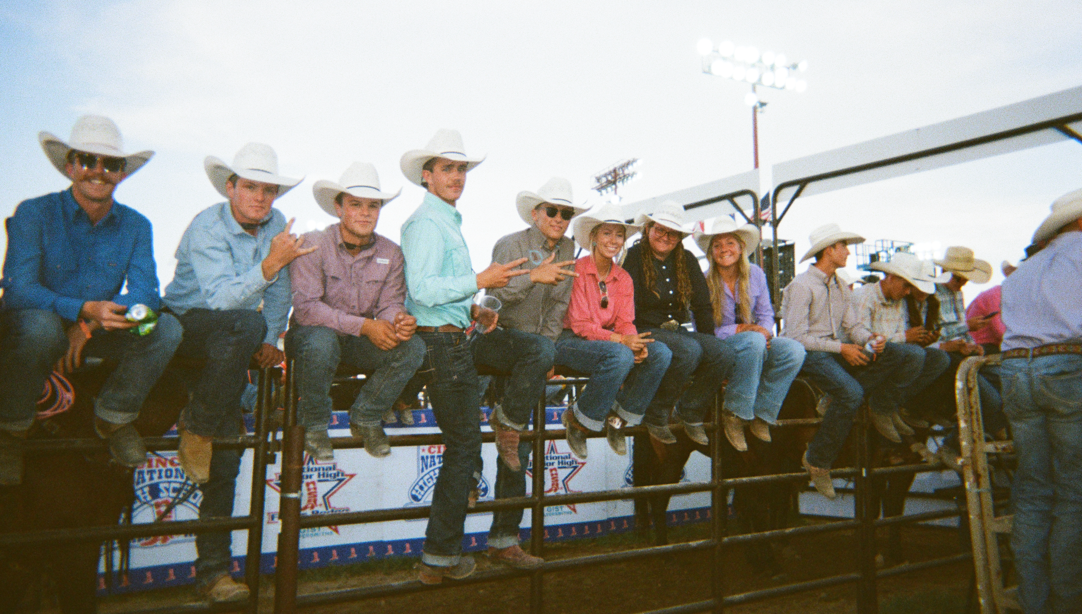 A group of people wearing cowboy hats and western clothing sit along a metal barrier at an outdoor rodeo or fair event under a bright sky.