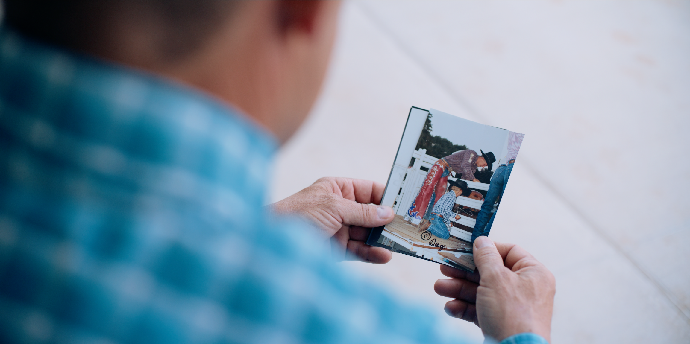 A person in a blue plaid shirt holds a small printed photograph or card in both hands, shown from behind over a light-colored floor.