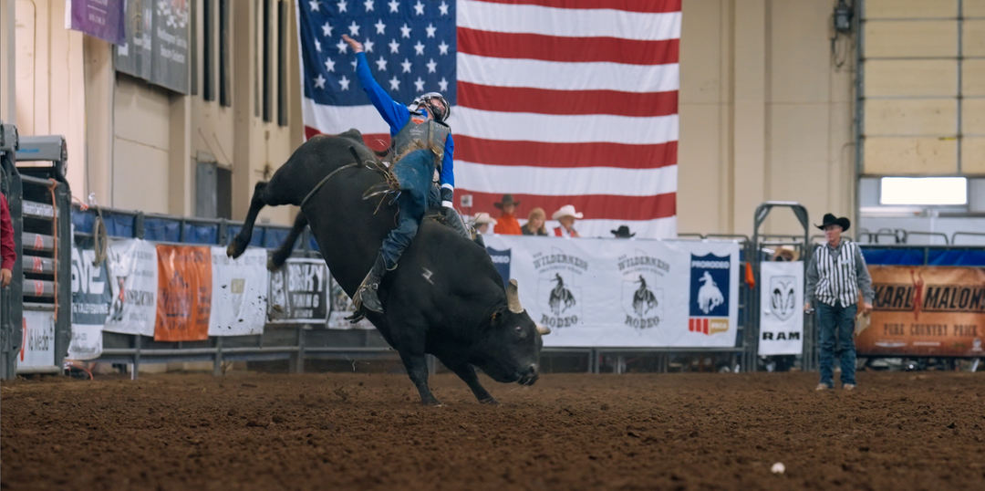 A cowboy in a blue shirt and jeans rides a bucking black bull in an indoor rodeo arena, with an American flag and event banners in the background.
