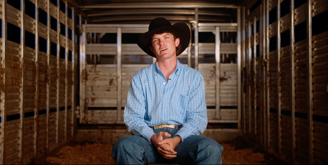 A man wearing a black cowboy hat, a light blue striped long-sleeve shirt, and blue jeans sits inside the aisle of a metal livestock trailer.