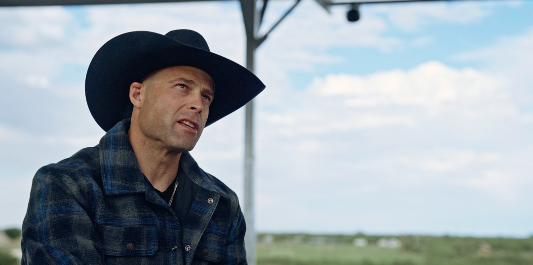 A man wearing a black cowboy hat and a dark blue plaid flannel shirt, looking off-camera under a covered outdoor structure with a cloudy sky in the background.