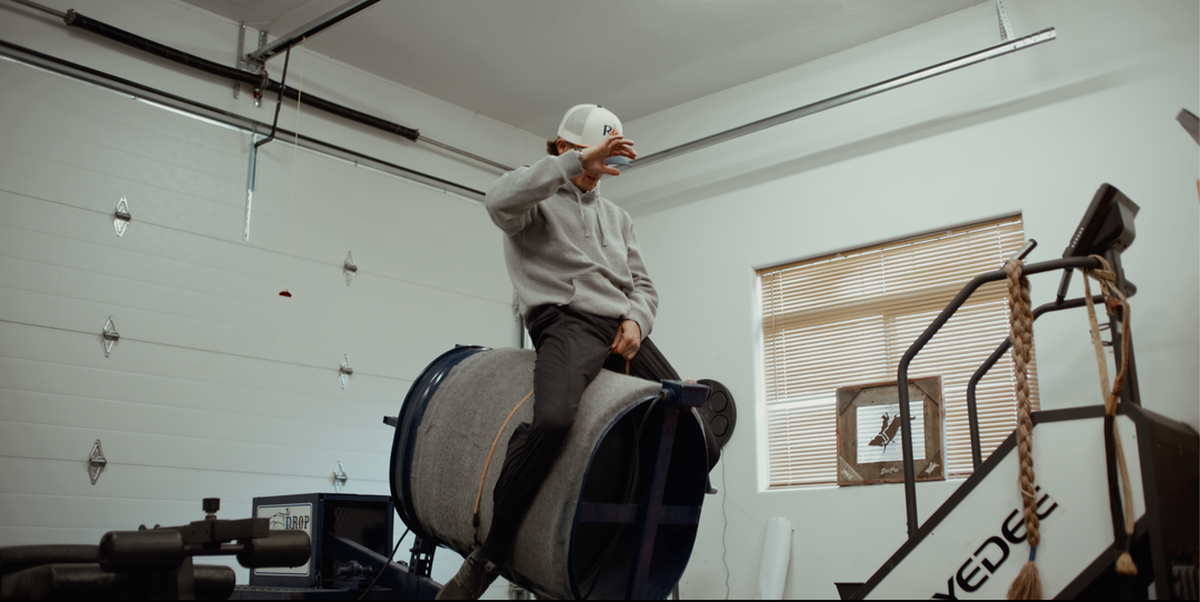 A man in a white hat and gray sweatshirt sits on a bull riding training machine inside a white garage-like workshop with a window and industrial equipment visible.