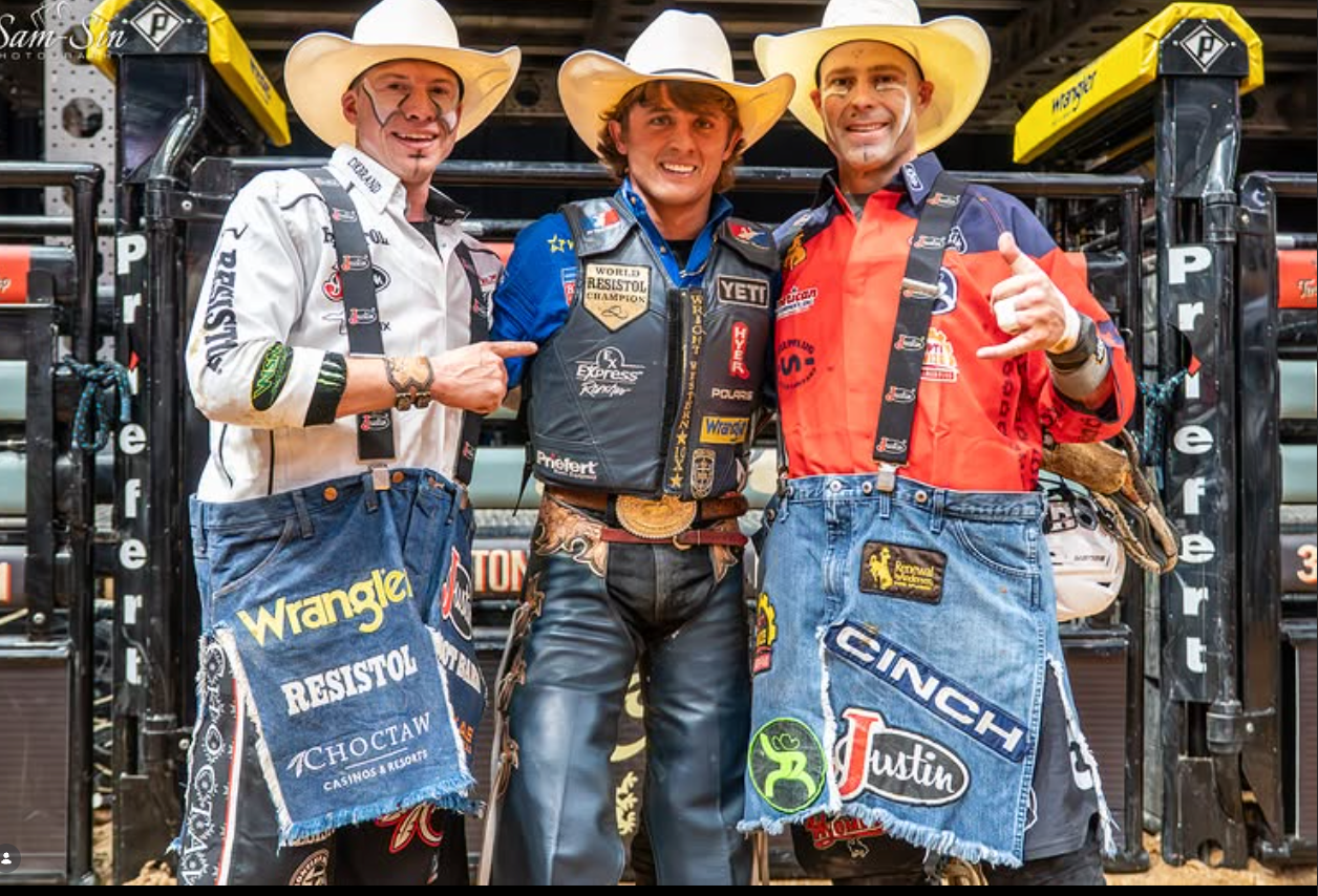 Three men in cowboy hats and bull riding gear pose together in front of a rodeo arena gate. 