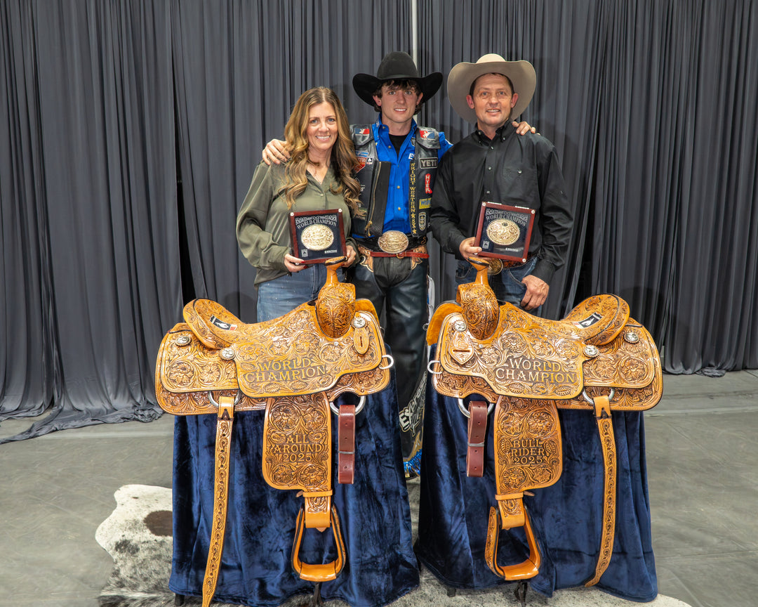 Three people in cowboy attire stand behind two ornate tooled Western saddles displayed on blue velvet stands, each holding a framed award or plaque, in front of a dark curtain backdrop.