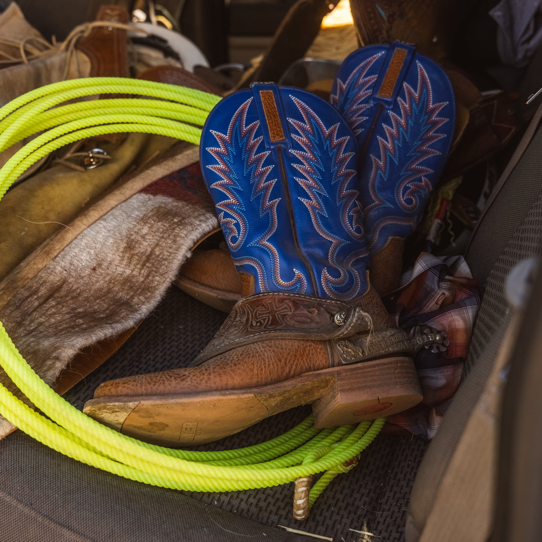 A pair of blue and brown western cowboy boots with intricate embroidery, resting on a dark textured surface next to a bright neon yellow rope.