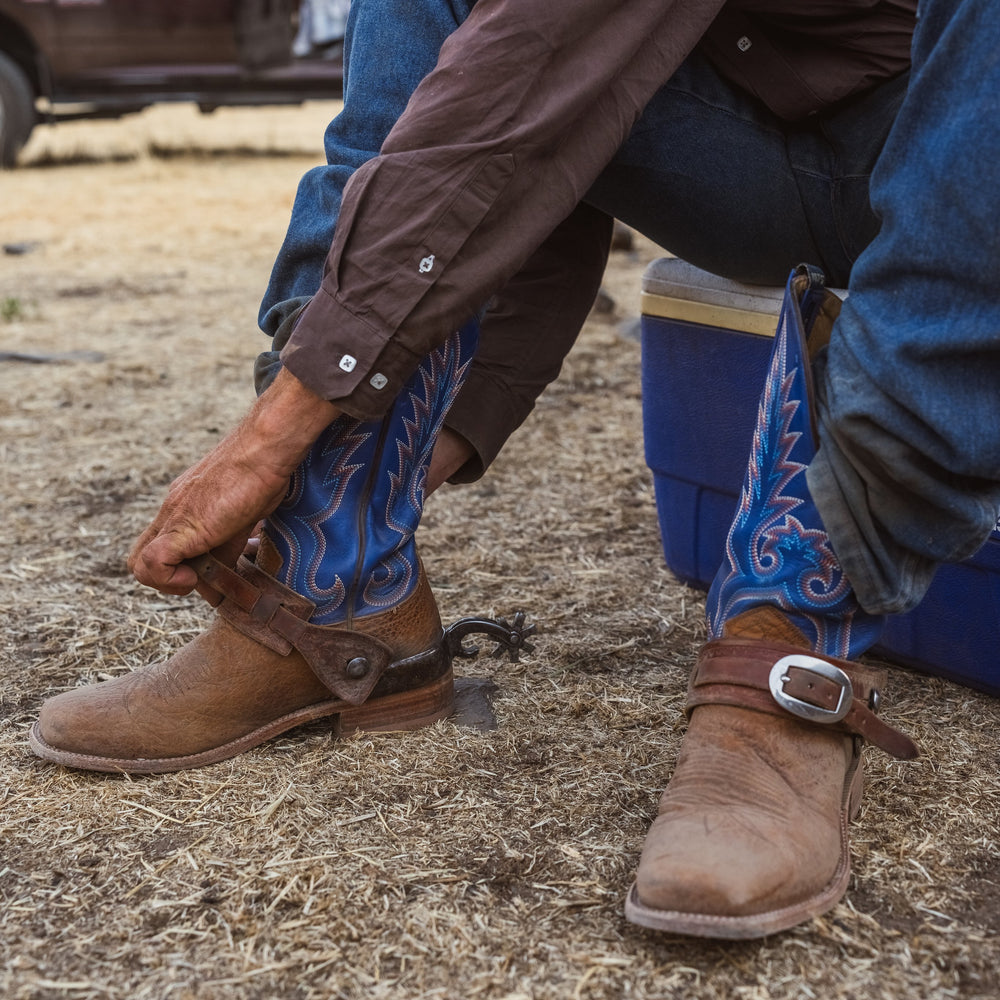 A close-up of a person sitting outdoors on a blue cooler, putting on a spurs with a strap and buckle while wearing blue and brown cowboy boots with embroidery.