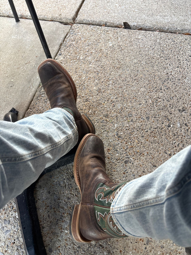 A person wearing brown leather cowboy boots with green and white western embroidery, paired with light-wash blue jeans, sitting on a black chair on a concrete sidewalk.