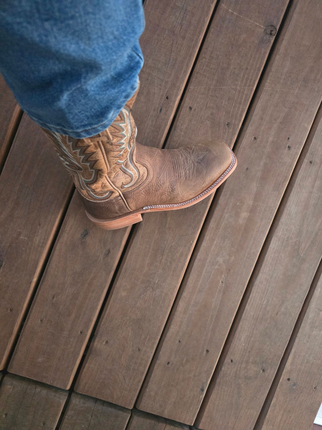A tan leather cowboy boot with western embroidery, worn with blue jeans on a brown wooden deck.
